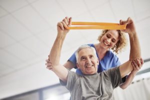 A senior man in a physical therapy session working with a band and assisted by a physical therapist.