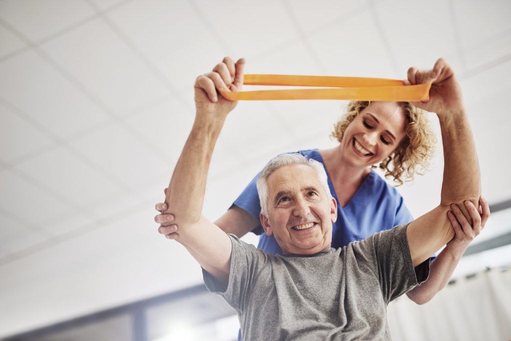 A senior man in a physical therapy session working with a band and assisted by a physical therapist.